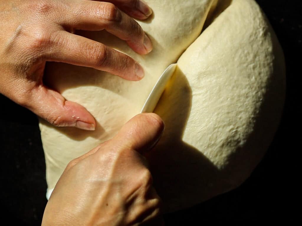 Hands seen from above knead and slice a large dough ball with a knife on a dark surface, highlighting the texture and action in natural light.